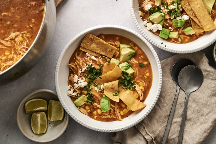 Top-down view of chicken tortilla soup served in white bowls, garnished with crispy tortilla strips, avocado, cilantro, and crumbled cheese, with lime wedges and a pot of soup in the background.