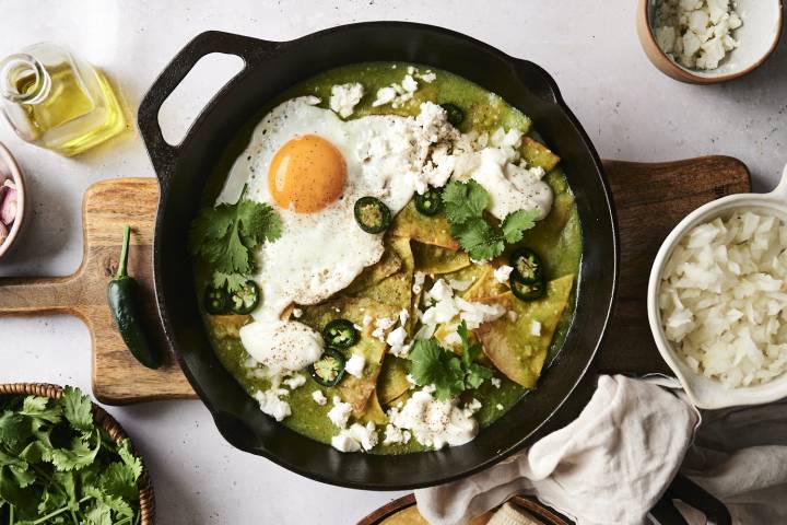 Chilaquiles verdes in a cast iron pan, topped with a fried egg, crumbled cheese, sour cream, jalapeño slices, and fresh cilantro. The pan is placed on a wooden board, surrounded by bowls of chopped onions, cheese, and fresh cilantro. A jalapeño and a bottle of oil are also nearby.