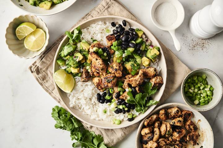 A bowl filled with grilled chili-lime chicken, white rice, black beans, avocado salsa, and fresh cilantro, garnished with lime wedges and green onions.