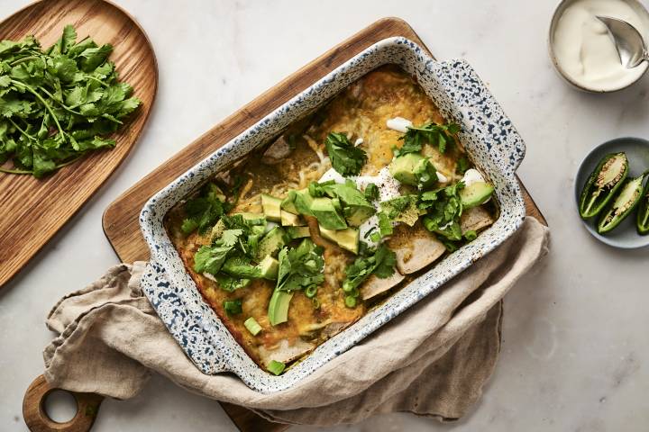 A baking dish filled with green chicken enchiladas, topped with melted cheese, avocado, cilantro, sour cream, and green onions, placed on a wooden cutting board with fresh cilantro and jalapeños on the side.