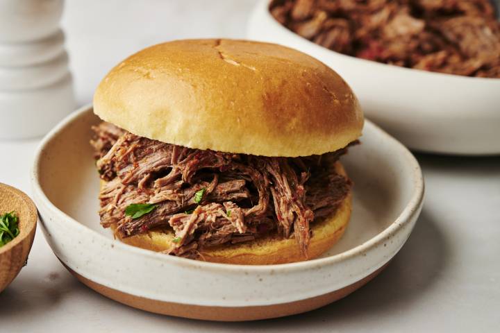 A shredded beef sandwich served on a soft golden bun, with tender, juicy beef and garnished with parsley, placed on a rustic ceramic plate