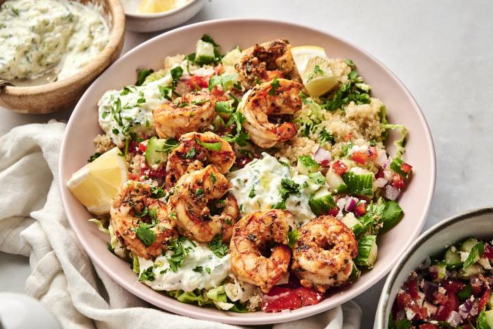 Mediterranean shrimp bowl featuring grilled shrimp, quinoa, chopped vegetables, tzatziki sauce, and lemon wedges, garnished with fresh parsley.