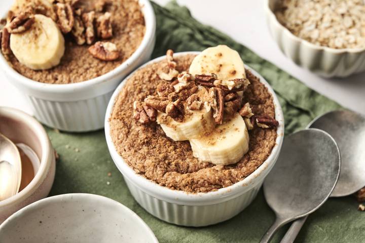 Two ramekins of baked protein oats topped with sliced bananas, chopped pecans, and a sprinkle of cinnamon, placed on a green napkin with a bowl of oats and spoons in the background.