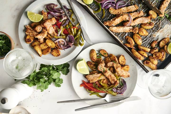 A sheet pan meal featuring seasoned pork chops, roasted potatoes, and sautéed bell peppers with red onions, served on plates with fresh cilantro and lime wedges.