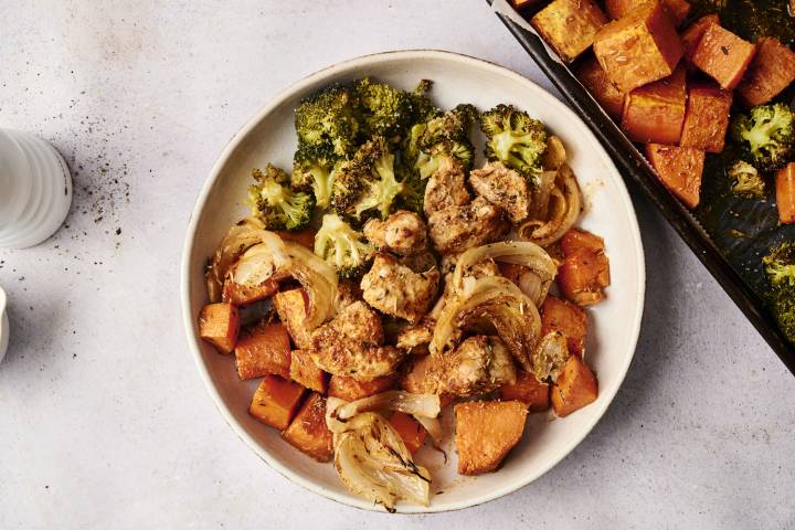 A serving of sheet pan herb chicken and sweet potatoes plated with roasted broccoli, caramelized onions, and seasoned sweet potato chunks. The plate is set on a light background with a portion of the sheet pan visible beside it.