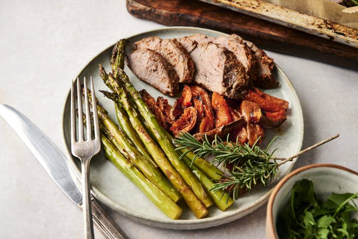 A close-up view of a dinner plate featuring juicy rosemary-seasoned steak slices, perfectly roasted asparagus, and golden sweet potato wedges, plated elegantly with a sprig of fresh rosemary and accompanied by a fork and knife