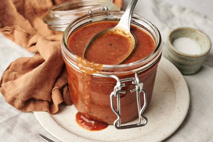 A glass jar filled with homemade enchilada sauce, with a spoon resting inside, placed on a ceramic plate with a rustic cloth and a small bowl of salt in the background.