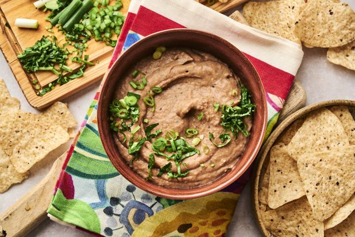 A bowl of creamy refried beans is garnished with chopped green onions and cilantro, served alongside tortilla chips on a colorful napkin.