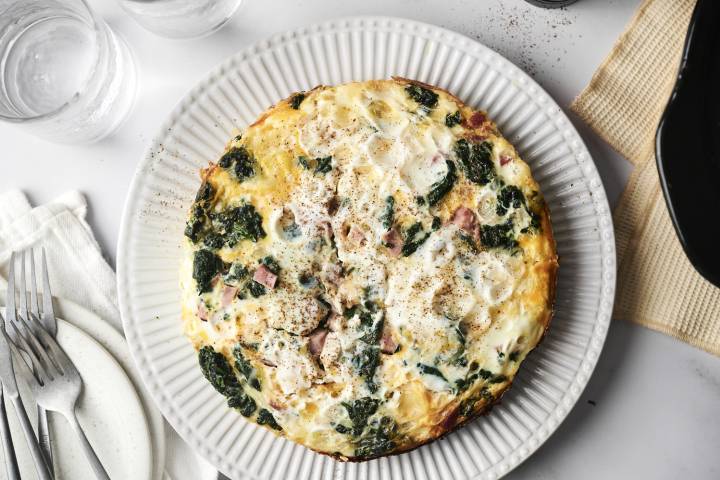 A close-up of a slice of breakfast casserole with layers of hash browns, eggs, ham, spinach, and cheese on a plate, with a fork resting beside it.