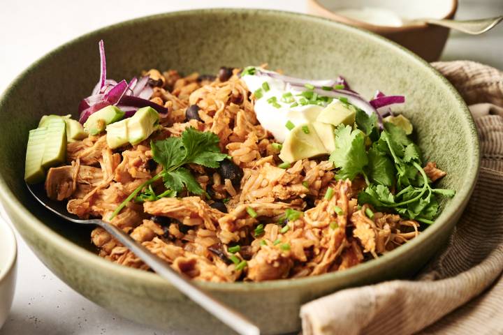 A chicken burrito bowl topped with avocado, cilantro, red onions, sour cream, and chopped chives, served in a green ceramic dish with a spoon.