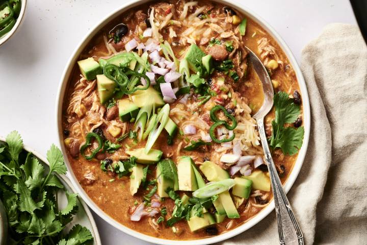 Slow cooker chicken enchilada soup in a white bowl, garnished with avocado, jalapeños, red onion, cilantro, green onions, and shredded cheese, served with a spoon.