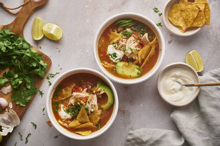 Two bowls of slow cooker chicken fajita soup topped with crispy tortilla chips, avocado slices, sour cream, and fresh cilantro, served with lime wedges and garnishes on a neutral background.