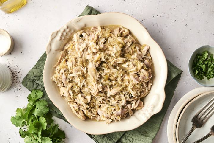 Slow-cooked shredded chicken in a tangy chile verde sauce, served in a rustic dish with cilantro garnish on the side.