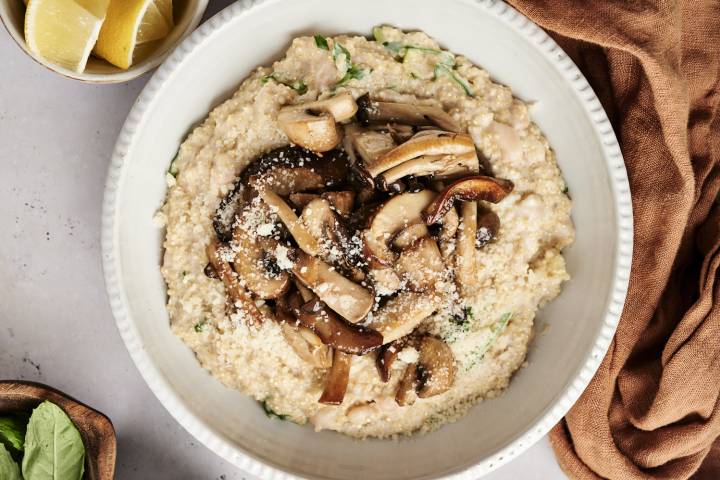 Creamy quinoa topped with sautéed mushrooms and Parmesan cheese, served in a white bowl with lemon wedges on the side.