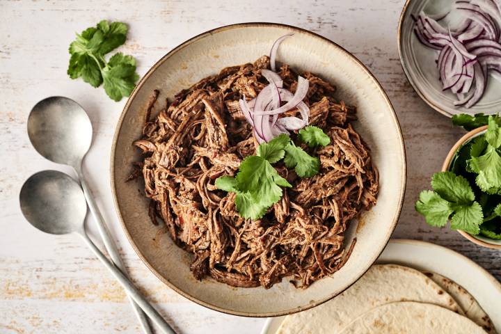 A bowl of tender, shredded Mexican beef adobo is garnished with fresh cilantro and sliced red onions, ready to be served with tortillas.