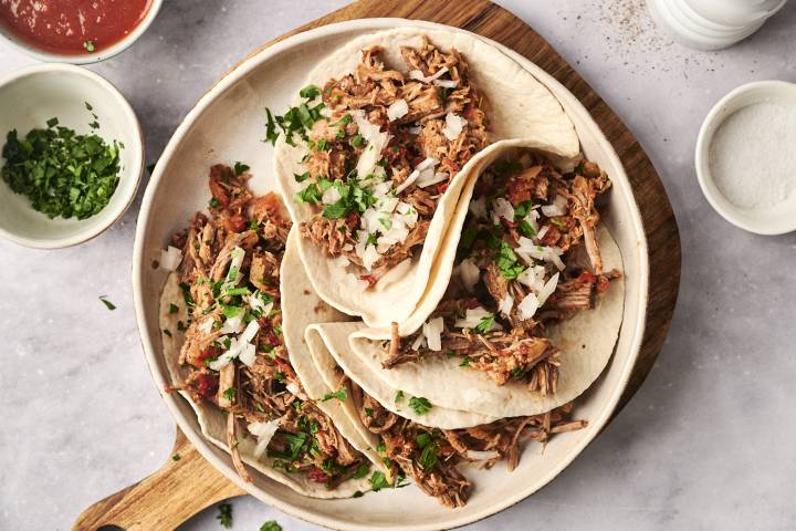 A plate of soft flour tortillas filled with slow-cooked spicy braised beef, topped with chopped onions and fresh cilantro, served with salsa on the side.