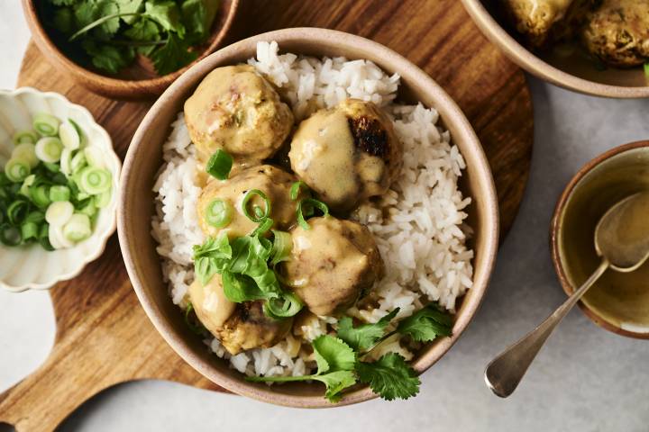 A bowl of white rice topped with Thai curry chicken meatballs, coated in a creamy sauce and garnished with fresh cilantro and sliced green onions.