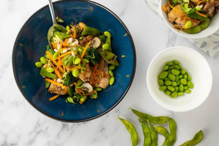 A vibrant dish of soy-glazed chicken and vegetables served in a blue bowl, featuring edamame, broccoli, carrots, mushrooms, and snap peas.