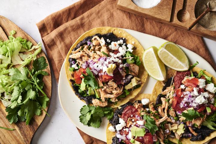 A  plate of tostadas, topped with black beans, shredded chicken, fresh lettuce, tomato, red onion, avocado, cilantro, and crumbled cheese. Lime wedges are placed on the side for a fresh squeeze of citrus.