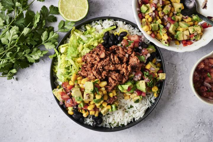 A vegan ground beef burrito bowl is served with cilantro lime rice, black beans, lettuce, seasoned plant-based ground beef, avocado, corn, and fresh pico de gallo, garnished with chopped cilantro and a lime wedge.