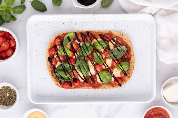 Baked Caprese flatbread on a baking sheet with balsamic glaze and fresh basil leaves.