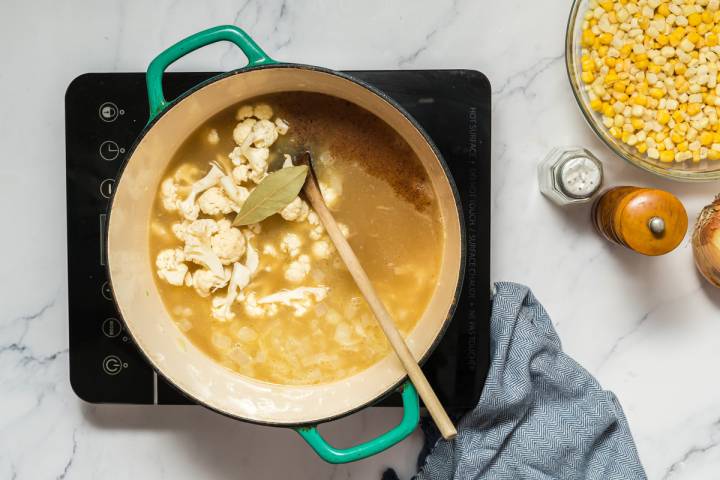 Cauliflower, vegetable broth, and spices being added to a soup pot for corn chowder.