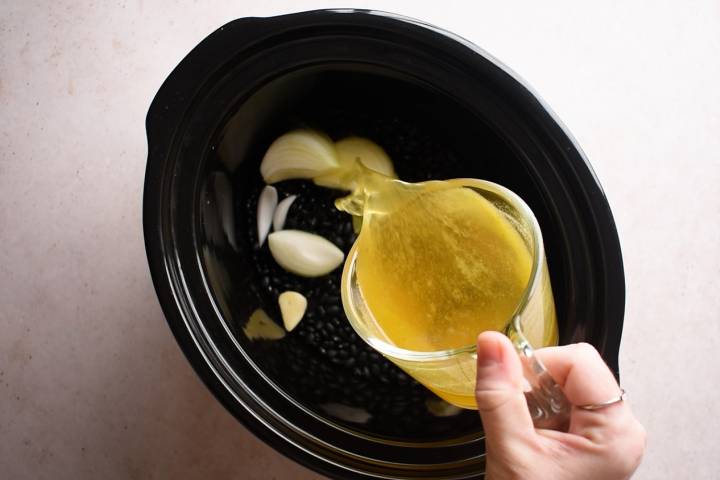 Vegetable broth being added to a slow cooker of black beans, onions, and garlic.