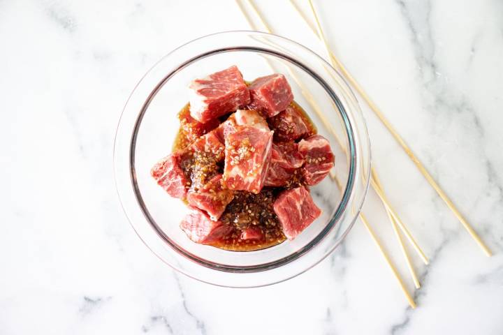 Cued steaks in a bowl with teriyaki sauce and wooden skewers on the side.