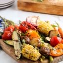 Grilled vegetables including zucchini, peppers, tomatoes, potatoes, and carrots on a wooden cutting board.