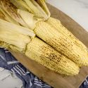 Grilled corn with the husks attached piled on a wood cutting board.