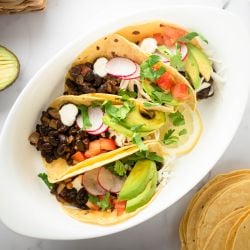 Black bean tacos with chipotle peppers served in corn tortillas topped with avocado, radish, tomatoes, and fresh cilantro.