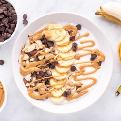 Chunky monkey banana peanut butter Greek yogurt bowl with sliced bananas, almonds, chocolate chips, and melted peanut butter.