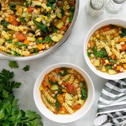 Minestrone soup with zucchini, yellow squash, pasta, white beans, spinach, and tomatoes in two bowls. 