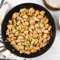 Healthy Sesame Chicken in a skillet with green onions with white rice and broccoli on the side.