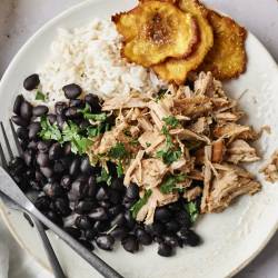 Shredded garlic Cuban pork served with black beans, rice, and crispy plantains on a white plate. The pork is topped with fresh cilantro, and there is a fork and knife on the side. 