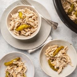 Slow cooker pepperoncini chicken shredded in three bowls with rice on the side and whole pepperoncini peppers.