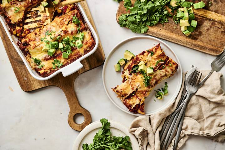 Cheesy black bean enchiladas topped with diced avocado and fresh cilantro, served on a white plate with a side of extra herbs on a wooden cutting board.