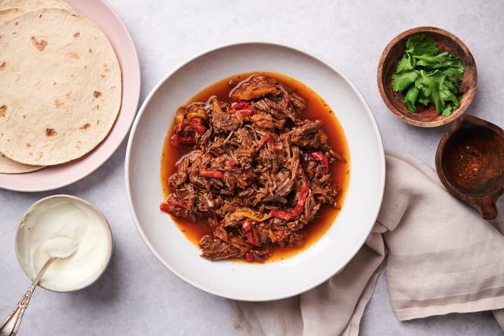 Crockpot beef fajitas with bell peppers, onions, and fajita sauce in a bowl with tortillas, cilantro, and sour cream on the side.