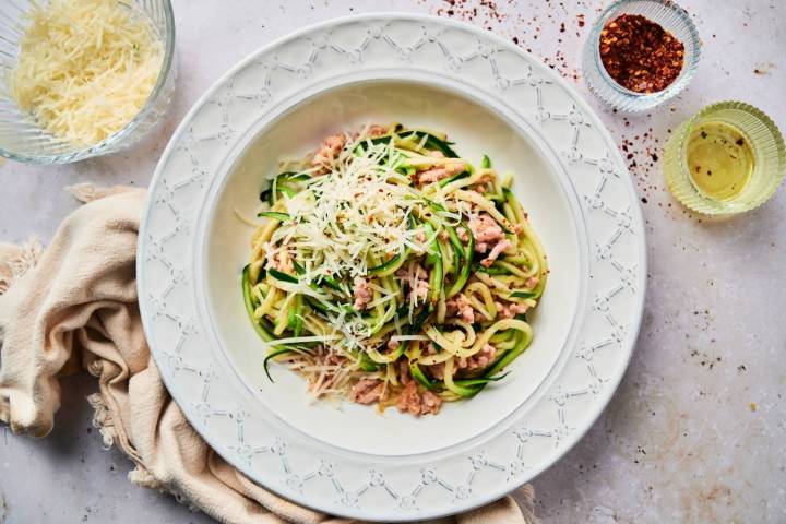 Garlic Parmesan zucchini noodles served in a white dish, topped with grated cheese and red pepper flakes, accompanied by ingredients like olive oil and shredded Parmesan cheese.