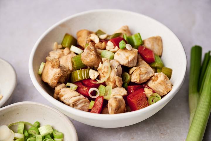 A bowl of Healthy Kung Pao Chicken featuring chunks of chicken, red bell peppers, celery, and garnished with green onions and peanuts.