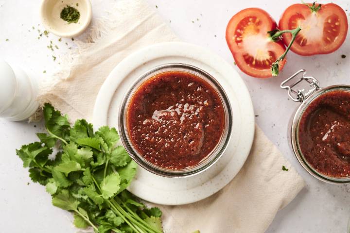 A jar of rich red enchilada sauce surrounded by fresh cilantro, tomatoes, and seasonings on a white backdrop.