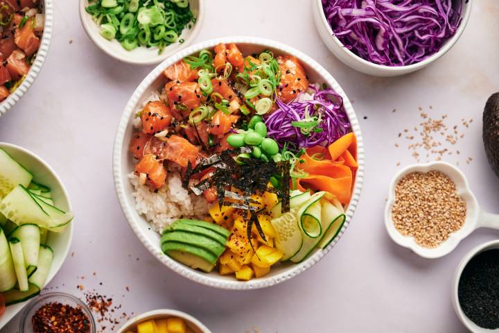 Salmon poke bowls with raw salmon, white rice, avocado, green onions, cucumber, seaweed, edamame, and cabbage.