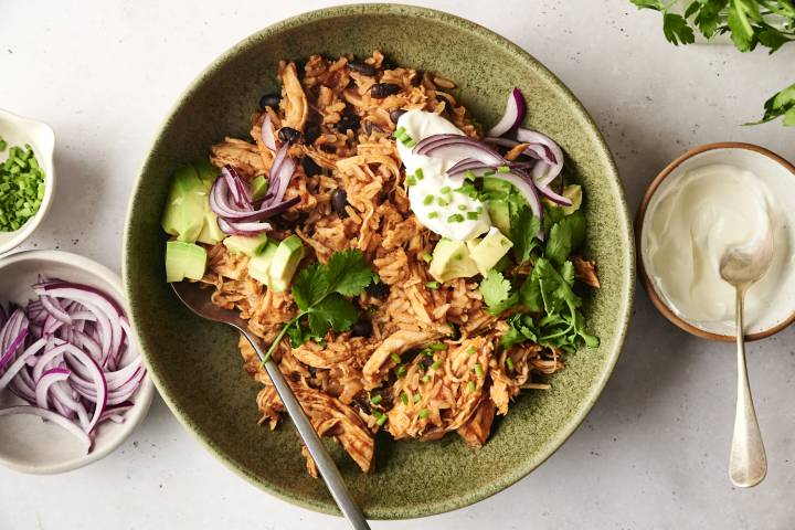 A chicken burrito bowl topped with avocado, cilantro, red onions, sour cream, and chopped chives, served in a green ceramic dish with a spoon.