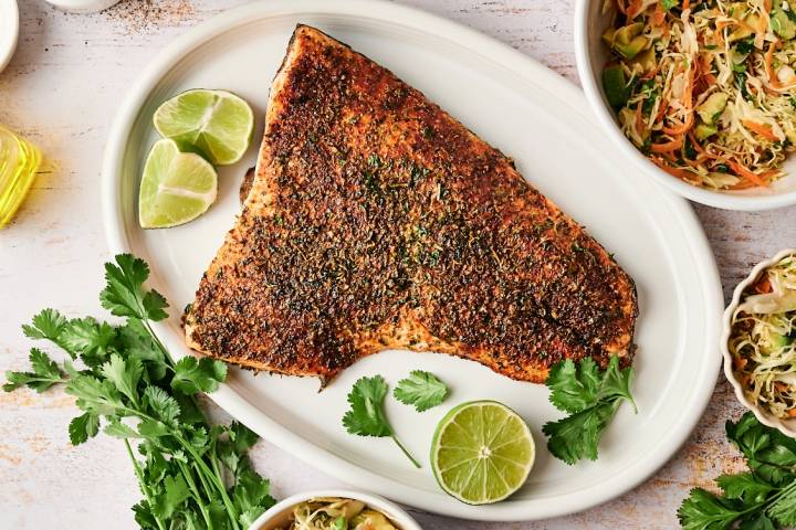 A whole taco spiced salmon fillet is served on a white platter with lime wedges, fresh cilantro, and bowls of slaw on the side.