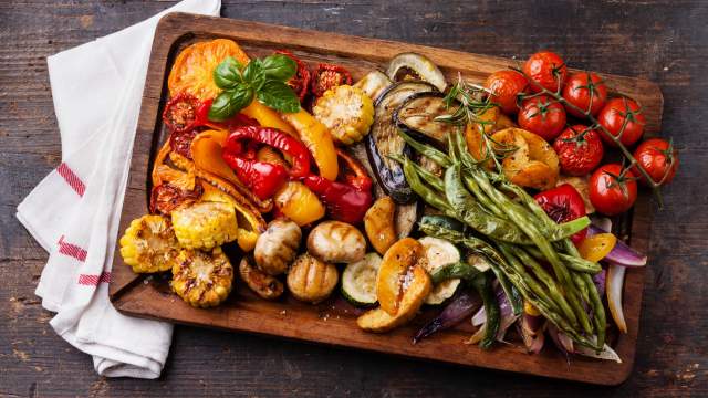 Grilled vegetables including peppers, tomatoes, asparagus, mushrooms, and corn on a wooden board.