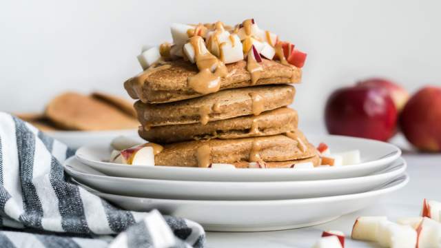 Apple cinnamon pancakes stacked on a plate with melted peanut butter and chopped apples.