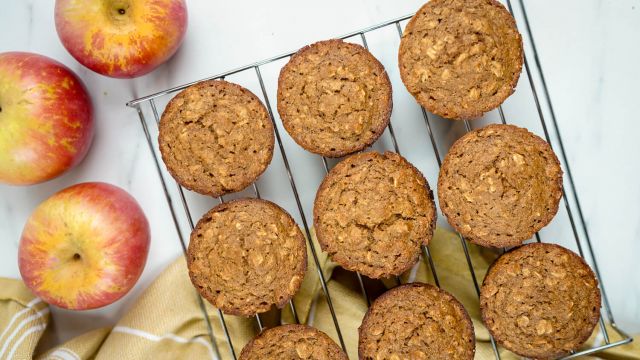 Applesauce oatmeal muffins with oatmeal flecks on a baking rack with a yellow napkin and apples.