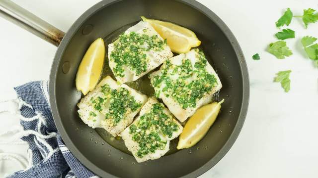 Baked cod with garlic, butter, lemon, and parsley with fresh lemon wedges in a small pan.