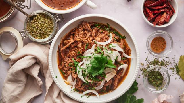 Birria de res with a red chile sauce served in a bowl with corn tortillas, sliced onions, cilantro, and consome.