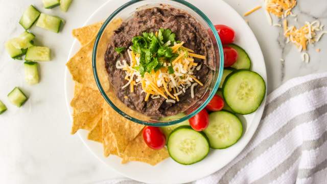 Easy black bean dip in a bowl with shredded cheese and cilantro with cucumbers, tomatoes, and chips on the side.